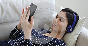 Asian woman in headphones lying on couch using smart phone