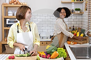 Asian woman and her African American friend is helping each other preparing healthy food with variety of vegetables while washing