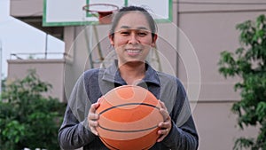 Asian woman holding basketball looking at camera at outdoor basketball playground.