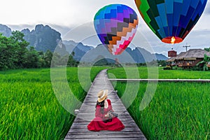 Asian woman holding a camera sits on a wooden walkway and looks at balloons with green rice fields in Vang Vieng, Laos