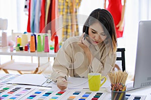 Asian woman holding hand smartphone talking with customer. Fashion designer clothes in the Tailor shop of fabrics store. Concept