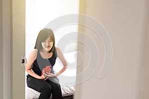 Asian woman holding hot water bag or bottle on her stomach ache sitting in bedroom