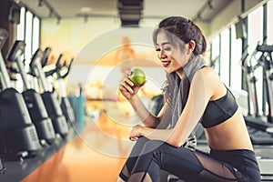 Asian woman holding and looking green apple to eat with sports equipment and treadmill in background. Clean food and Healthy