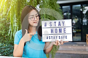 Asian woman holding and pointing a lightbox sign with text hashtag #STAY HOME and #STAY SAFE. COVID-19. Stay home save concept