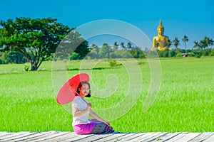 An Asian woman holding a red umbrella sitting on wooden bridge in a rice field with a large golden Buddha image