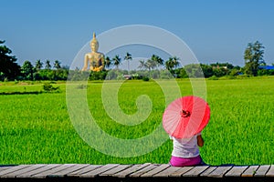 Asian woman holding a red umbrella sitting on a wooden bridge in a rice field with a large golden Buddha image