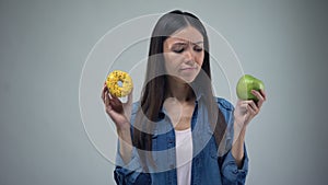 Asian woman holding sweet greasy donut and juicy green apple in hands, decision