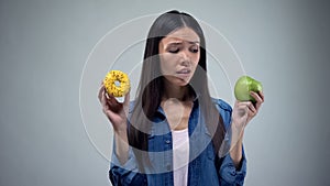 Asian woman holding sweet greasy donut and juicy green apple in hands, decision