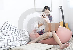 An Asian woman is holding a white maltese dog and using a laptop in the living room where a guitar is placed
