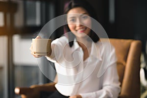 An Asian woman holds a cup of coffee and gives it to invite to drink together.Focus on the coffee cup