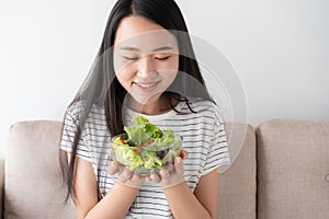 Asian woman holds a glass bowl full of salad vegetables green while sitting on the sofa at home. healthy and lifestyle concept