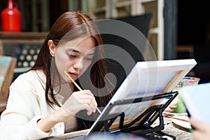 Asian woman holds a paintbrush in right hand as dominant hand, left hand is on laptop previewing illustration while drawing along