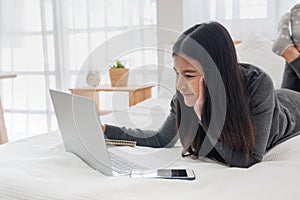 Asian woman laying on bed doing paperwork on laptop in bedroom.work at home.e-larning concept.study online photo