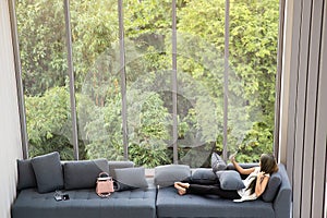 Asian woman laying on sofa near big glass wondows, relaxing alone in house with green forest in background