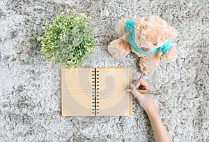 Asian woman lied on gray carpet textured background for write at the brown note book by a pen with artificial plant and bear doll