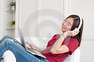 Asian woman listening music on headphone and using laptop computer with relax pose at couch in living room