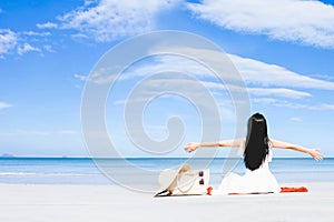 Asian woman, long black hair, wore white dress and hat sitting on the beach near the woven bags and sunglasses and facing back by