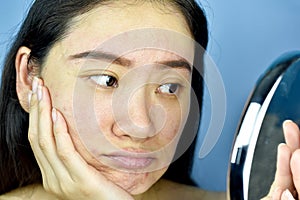 Asian woman looking at herself in the mirror, Female feeling annoy about her reflection appearance show the aging facial skin sign