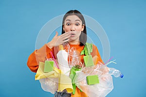 Asian woman looks shocked, holds plastic rubbish, sorting garbage for recycling, looks surprised, blue background