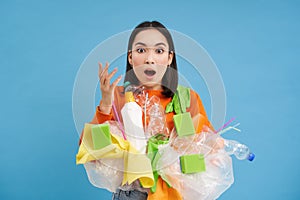 Asian woman looks shocked, holds plastic rubbish, sorting garbage for recycling, looks surprised, blue background