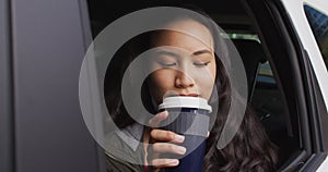 Asian woman with lowered face mask looking out of the window and drinking coffee sitting in the car