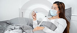 Asian woman in medical face mask feeling sick and holding thermometer to checking measure body temperature
