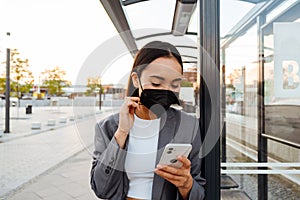 Asian woman in medical mask using mobile phone while standing at bus station