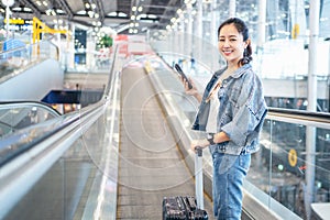 Asian woman with mobile phone in hand and luggage standing on escalator traveler at international airport, young girl tourist