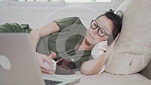 Asian woman mother working from home on computer laptop and feeding breast milk to baby infant. parent multitasking on sofa couch
