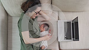 Asian woman mother working from home on computer laptop and feeding breast milk to baby infant. parent multitasking on sofa couch