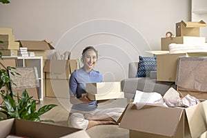 Asian woman moving into new apartment holding cardboard boxes with belongings, smiling and looking at camera