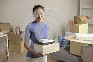 Asian woman moving into new apartment holding cardboard boxes with belongings, smiling and looking at camera