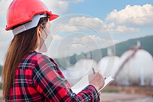 Asian woman petrochemical engineer wearing safety helmet standing holding a clipboard with checking gas storage industry and oil