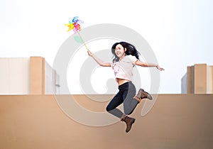 Asian woman posts, play and JUMPS with Windmill Toy colourful rainbow colour on the building rooftop in sunset time