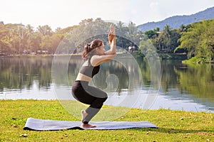 Asian woman practicing yoga doing Eagle Pose pose on the mat beside a lake in outdoor park