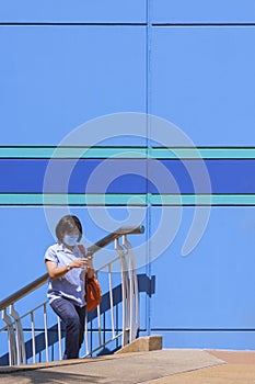 Asian woman in protective mask using smartphone while walking up the stairs outside of colorful department store