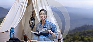 Asian woman reading book while on a solo trekking camp on the top of the mountain with small tent for weekend activities and