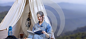 Asian woman reading book while on a solo trekking camp on the top of the mountain with small tent for weekend activities and