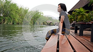 Asian woman relaxes by the river sitting on the edge of a wooden jetty