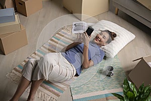 Asian woman relaxing at new house lying on floor using smartphone after moving in