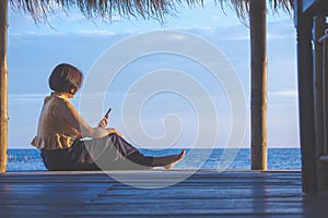 Asian woman is relaxing and using smartphone in cottage on seaside against seascape view background