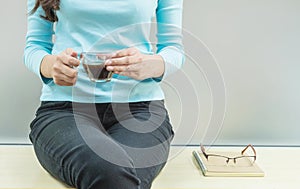 Asian woman rest for drink coffee in transparent glass cup in her hand in her free time in the room with wooden desk and frosted