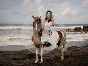 Asian woman riding horse on the beach. Outdoor activities. Woman wearing long white dress. Traveling concept. Cloudy sky. Copy