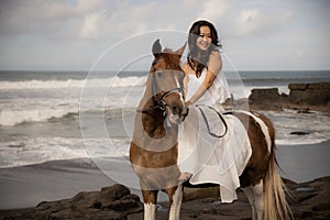 Asian woman riding horse on the beach. Outdoor activities. Woman wearing long white dress. Traveling concept. Cloudy sky. Copy