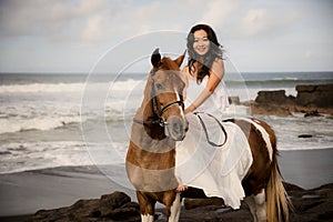 Asian woman riding horse on the beach. Outdoor activities. Woman wearing long white dress. Traveling concept. Cloudy sky. Copy