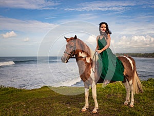Asian woman riding horse near the ocean. Outdoor activities. Woman wearing long green dress. Traveling concept. Cloudy sky. Copy