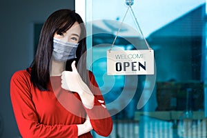 Asian woman shop owner smiling wearing medical face mask hanging label `Welcom we are open` and showing thump up. She open her s