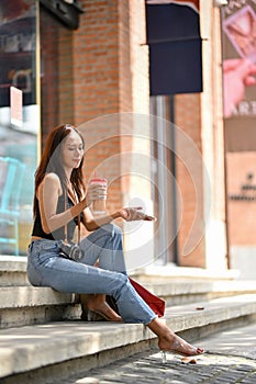 Asian woman with shopping bags, retro camera, coffee to go and smartphone in hand sitting on stairs on street mall