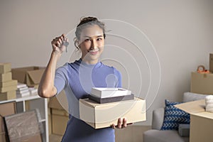 Asian woman showing keys and holding cardboard boxes in new house, smiling and looking at camera