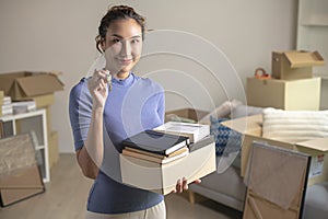 Asian woman showing keys and holding cardboard boxes in new house, smiling and looking at camera